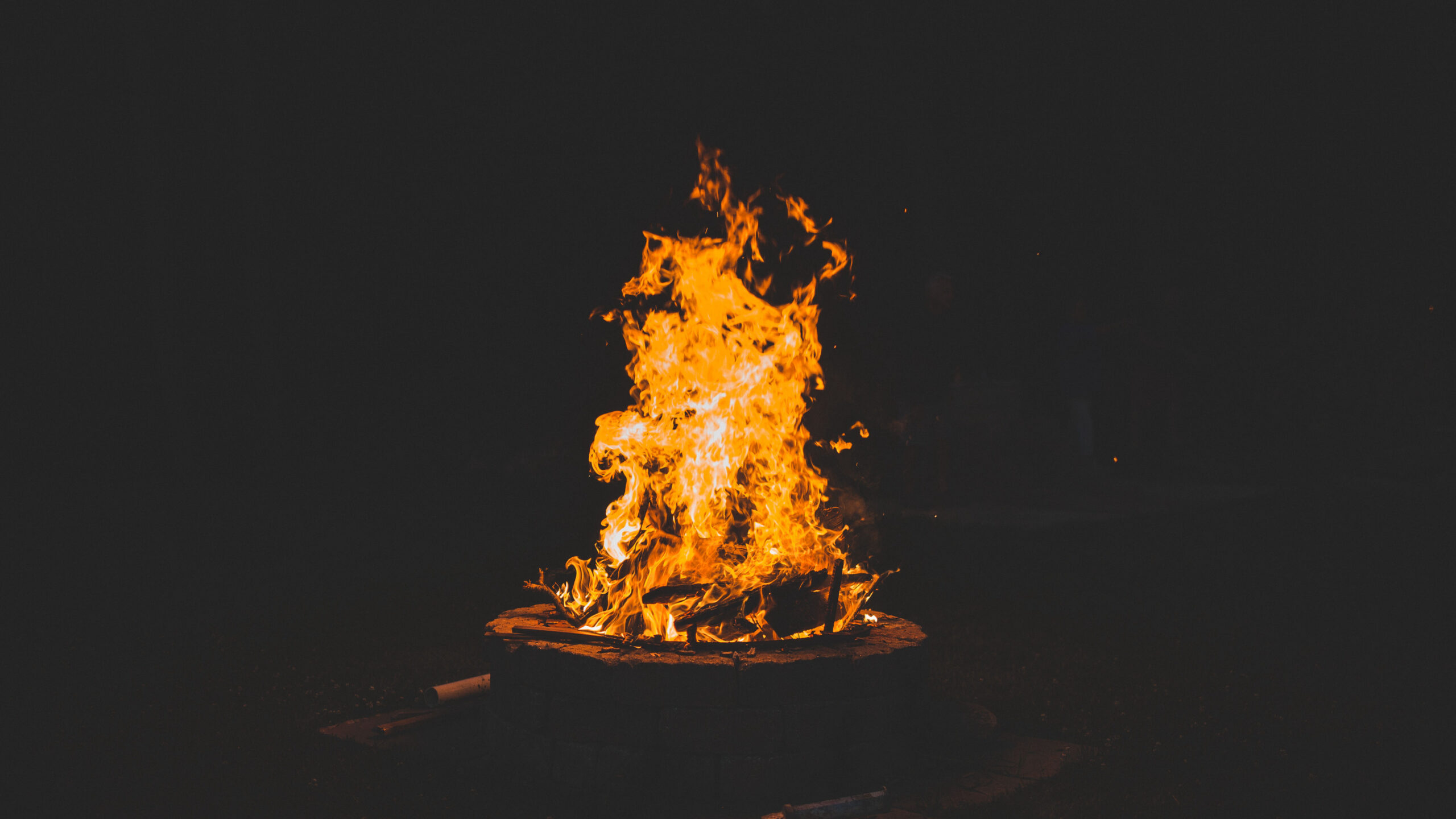 夜晚的户外,一个石制火盆里篝火正熊熊燃烧,橙黄色的火焰在漆黑的背景中向上跳动,散发着温暖的光芒。