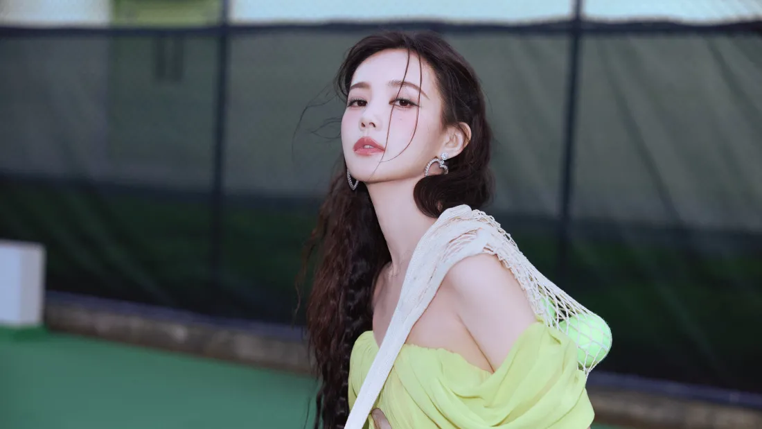 李一桐身着明黄色斜肩网球服,肩背白色网兜立于绿茵场,长发飘动的活力场景